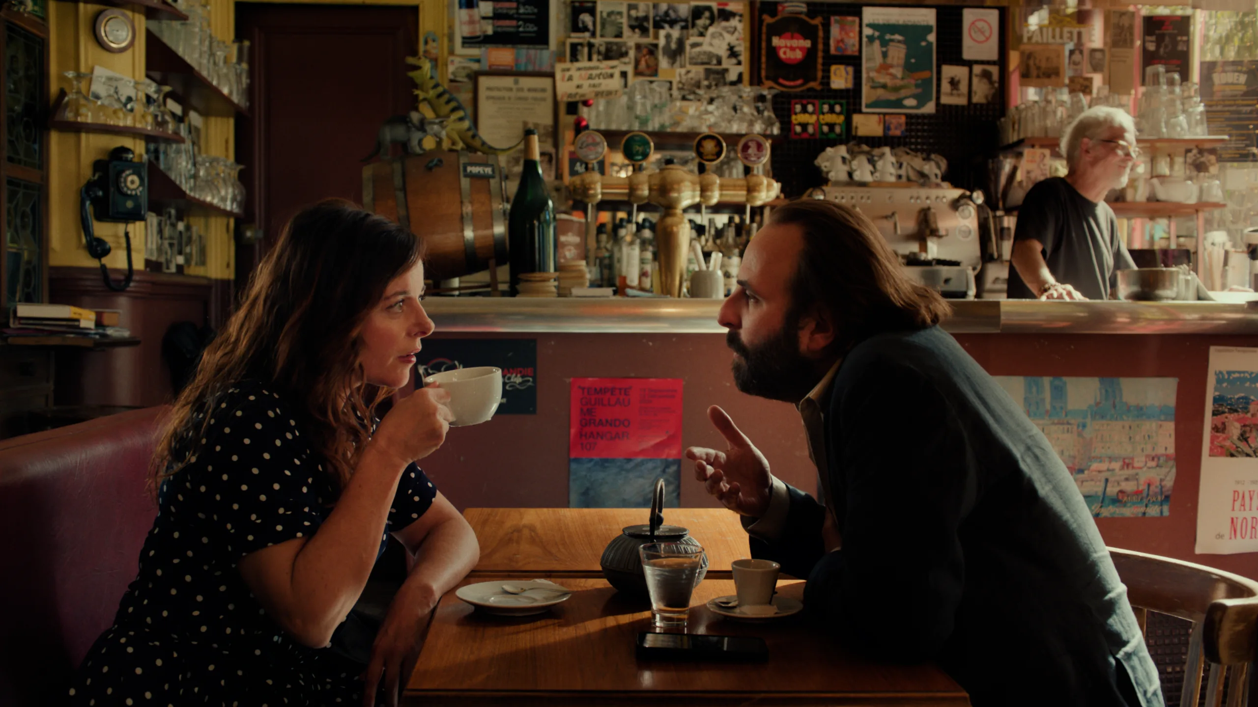 uma mulher está a beber café e um homem, à sua frente, fala com ela, estão num café