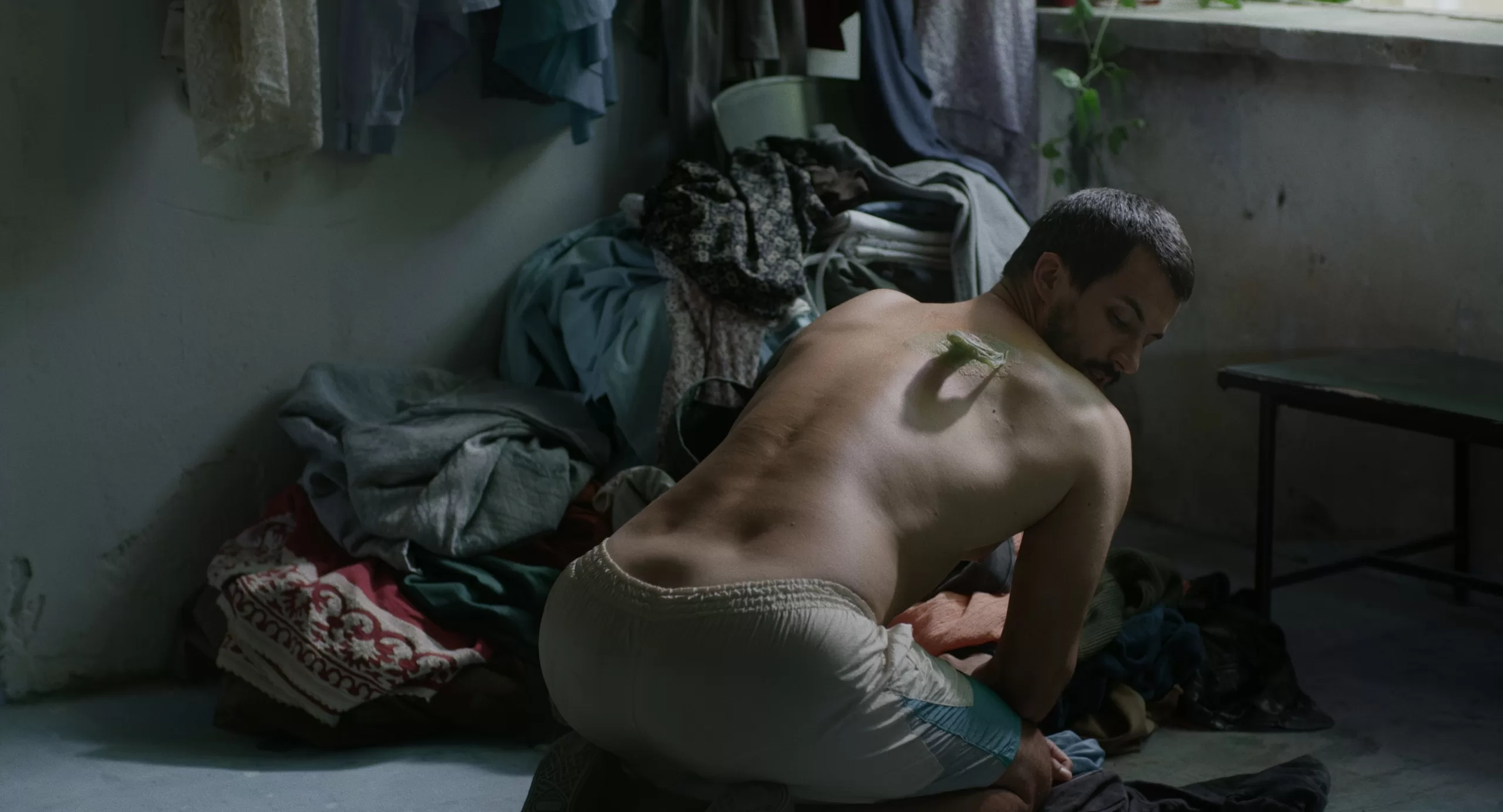 monte de roupa ao fundo, e um homem de joelhos, de costas, sem camisola, e com uma fita colada