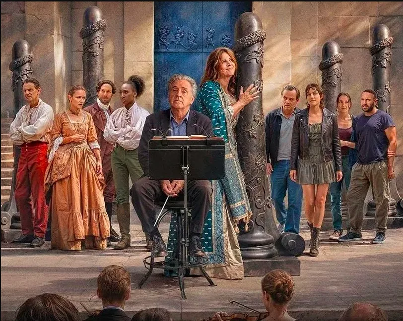 Um grupo de atores vestidos com vestuário de época e roupas modernas posa num cenário teatral decorado com colunas esculpidas com relevos de serpentes. No centro, um homem idoso sentado perante uma estante de partitura segura uma batuta, enquanto uma mulher ao seu lado segura uma coluna, sorrindo para cima. Em primeiro plano, vê-se a parte de trás da cabeça dos músicos de uma orquestra.