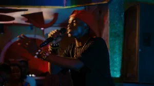 homem com chapéu para trás e microfone na mão, a cantar em cima de um palco