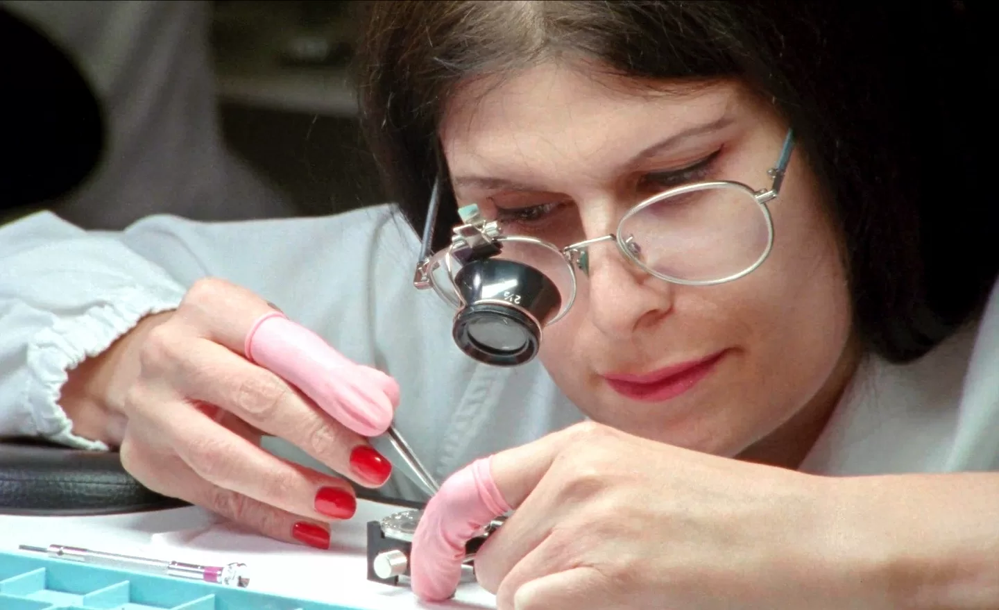 mulher a trabalhar com uma pinça, observando o objeto com uma lupa, e com luvas