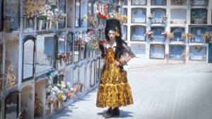 homem com um ar chateado, usa um vestido, segura um ramo de flores, e está no meio de um cemitério